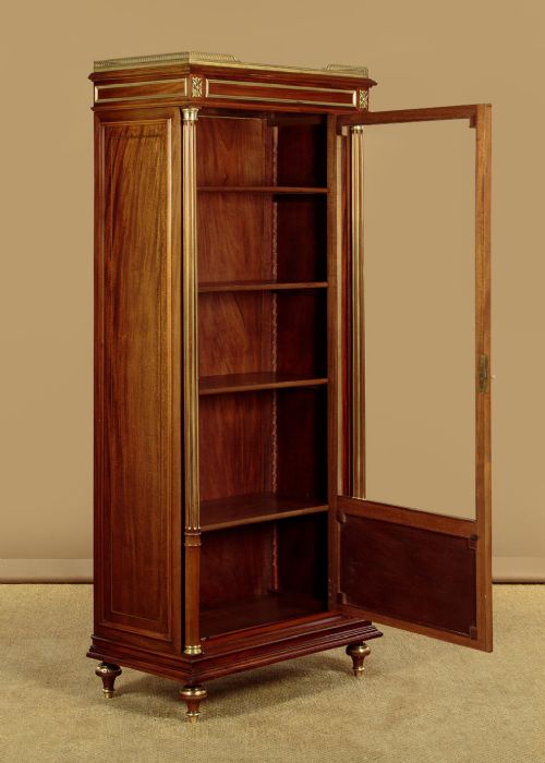 A tall, narrow mahogany bookcase featuring a single glazed door, internal wooden shelves, brass fluted column accents, and a brass gallery top. View V