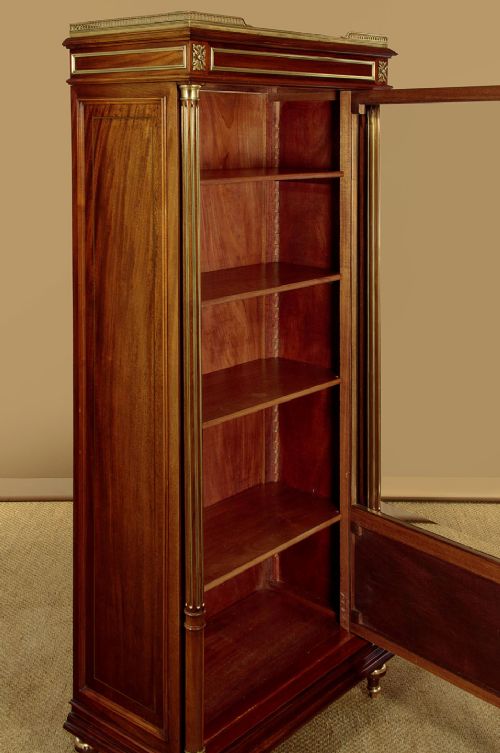 A tall, narrow mahogany bookcase featuring a single glazed door, internal wooden shelves, brass fluted column accents, and a brass gallery top. View X