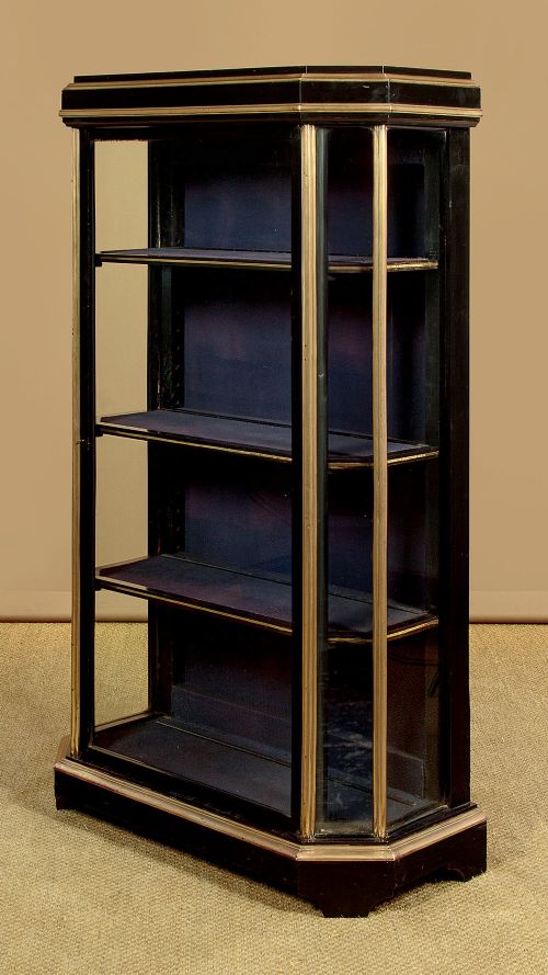 An Art Deco ebonised tall display cabinet featuring brass mounts, glazed side panels, a single glass door, and fabric-lined interior shelving. View V