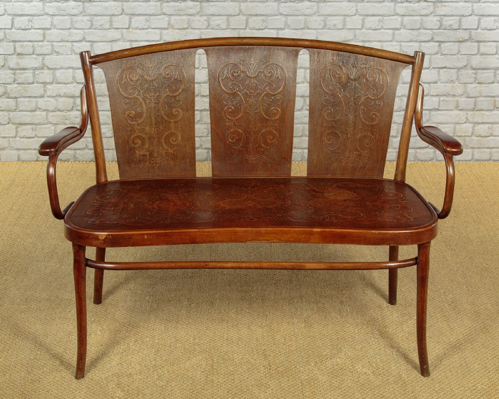 An antique bentwood settee with three embossed back panels, matching wooden seat, swept armrests, and a dark warm brown wood finish.