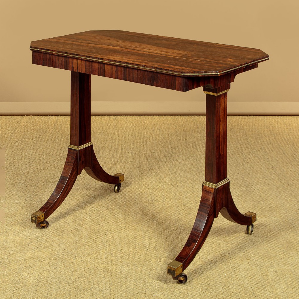 A Regency period rosewood writing table featuring a rectangular top with canted corners, brass beaded trim, twin end supports, and splayed legs with brass castors.