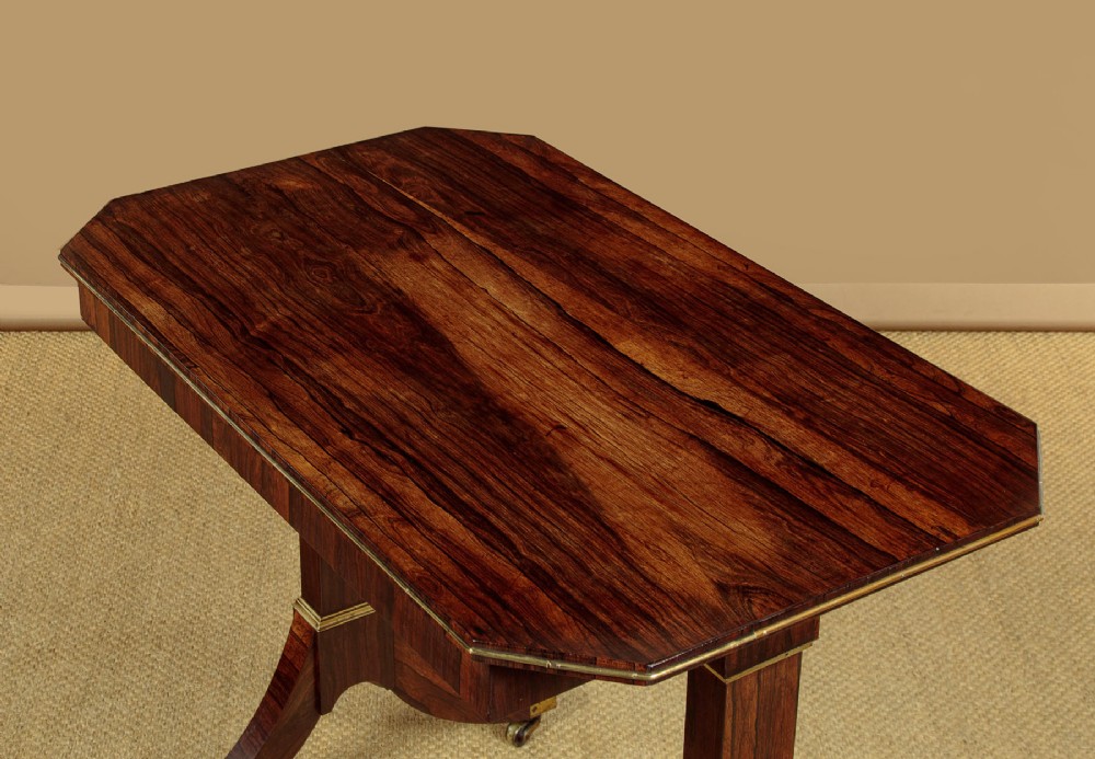 A Regency period rosewood writing table featuring a rectangular top with canted corners, brass beaded trim, twin end supports, and splayed legs with brass castors. View V