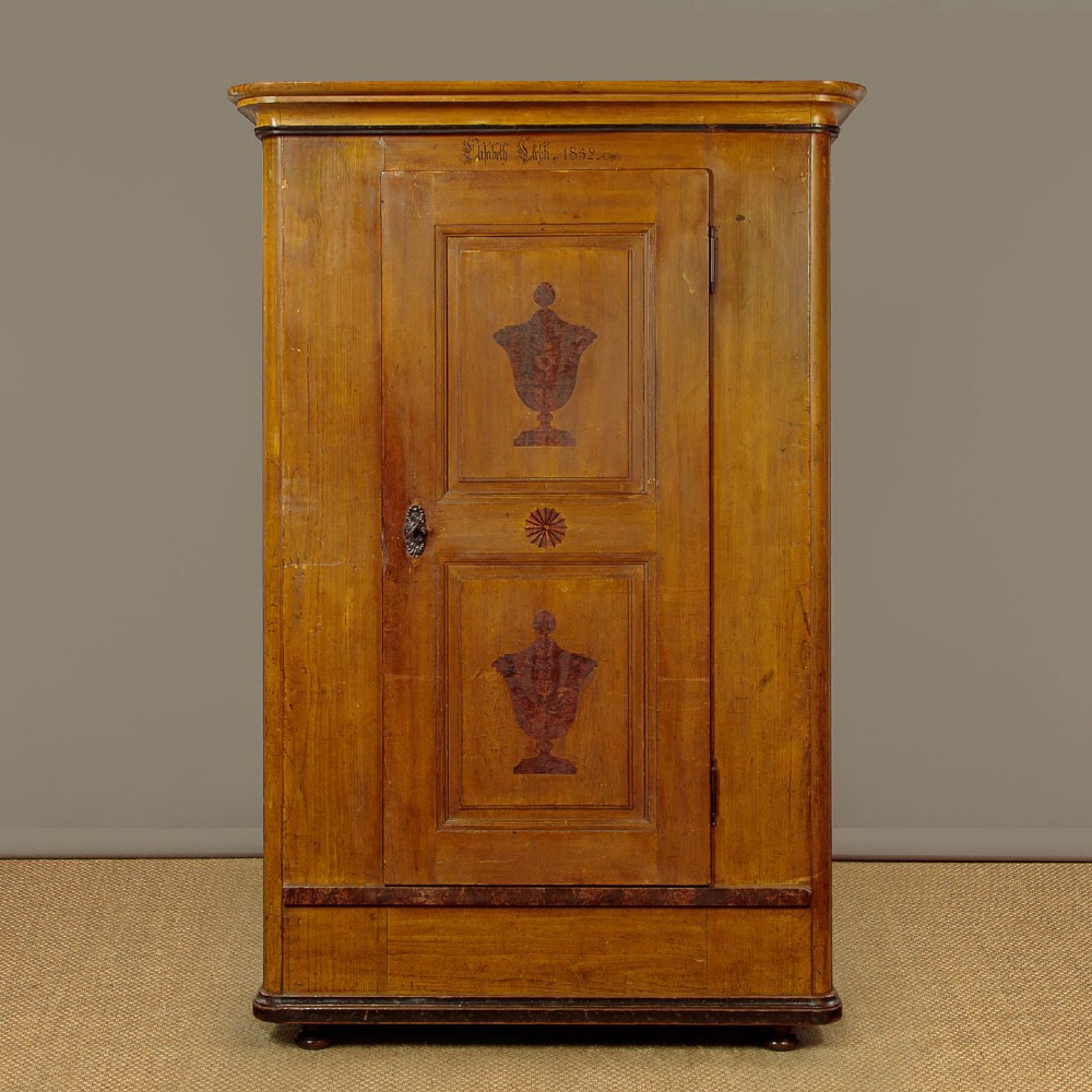 A single-door pine wardrobe featuring hand-painted urn motifs on two panelled sections and a dated inscription near the moulded cornice.