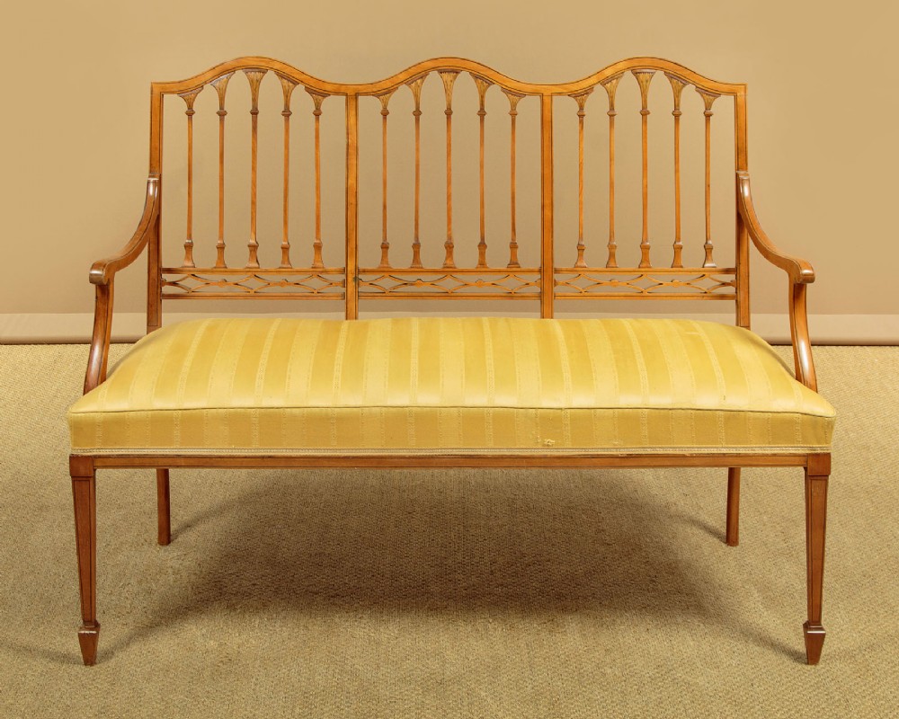 An antique inlaid satinwood triple chair-back settee featuring vertical splats, sweeping arms, and a yellow striped upholstered seat.