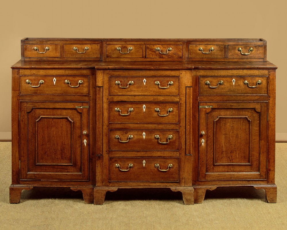 A late 18th-century warm brown oak Welsh dresser base featuring elevated top spice drawers, brass swan-neck handles, central drawers, and side cupboard doors.
