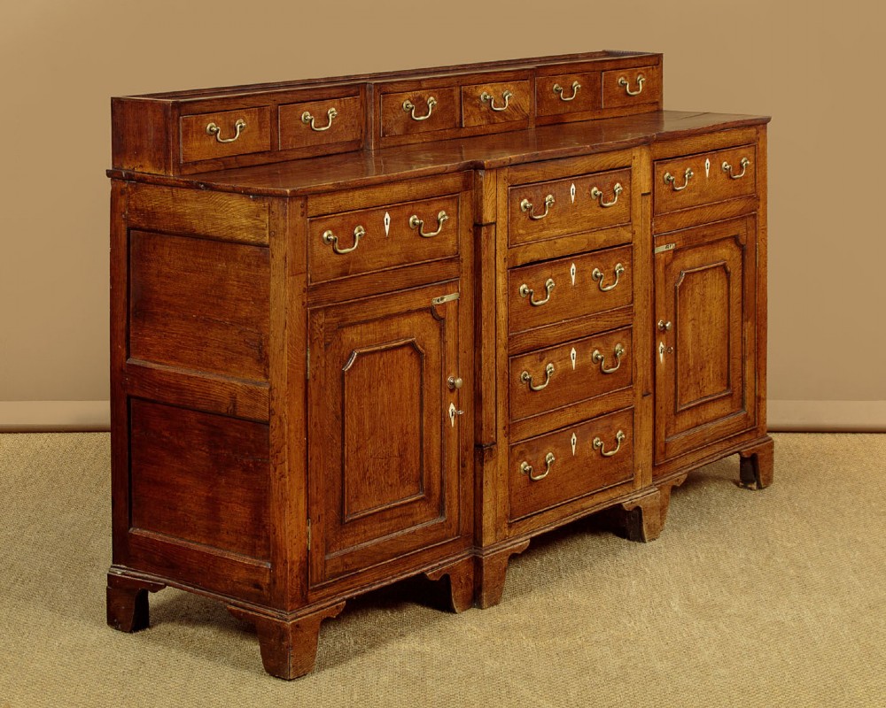 A late 18th-century warm brown oak Welsh dresser base featuring elevated top spice drawers, brass swan-neck handles, central drawers, and side cupboard doors. View III