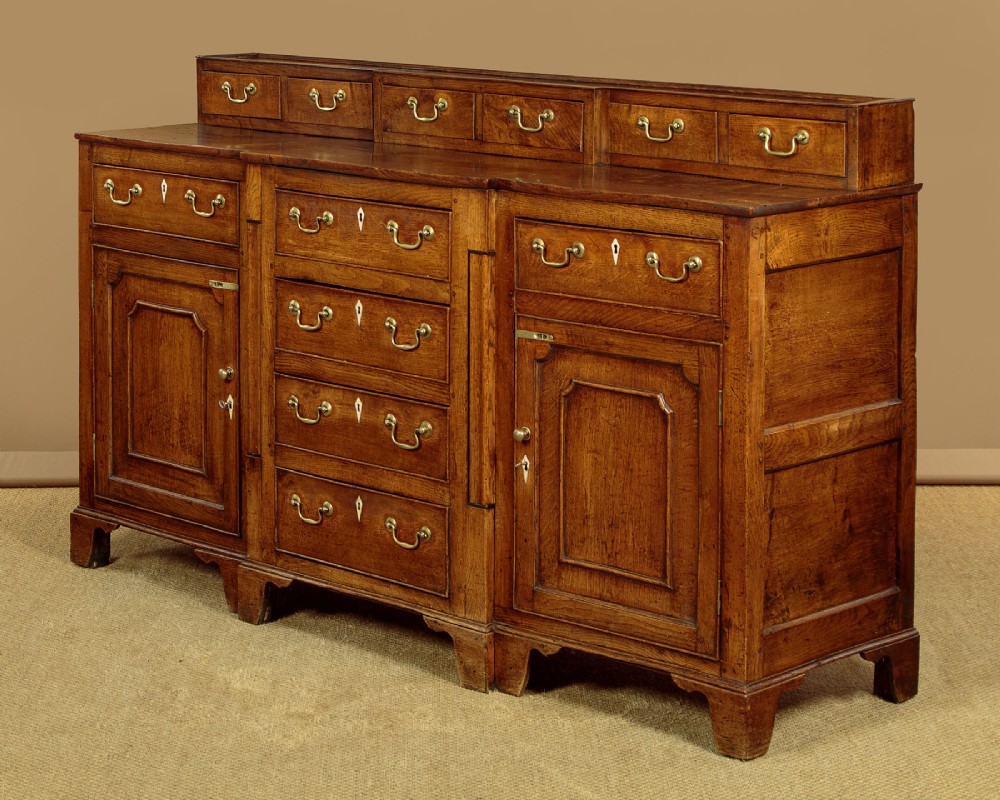 A late 18th-century warm brown oak Welsh dresser base featuring elevated top spice drawers, brass swan-neck handles, central drawers, and side cupboard doors. View IV