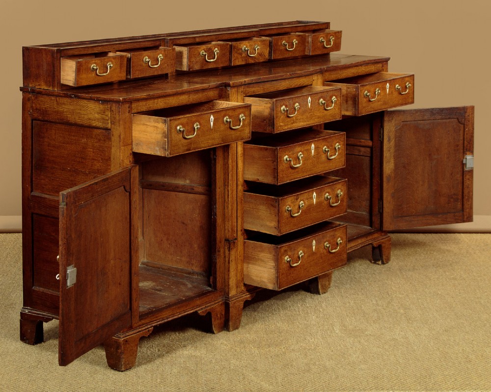 A late 18th-century warm brown oak Welsh dresser base featuring elevated top spice drawers, brass swan-neck handles, central drawers, and side cupboard doors. View V