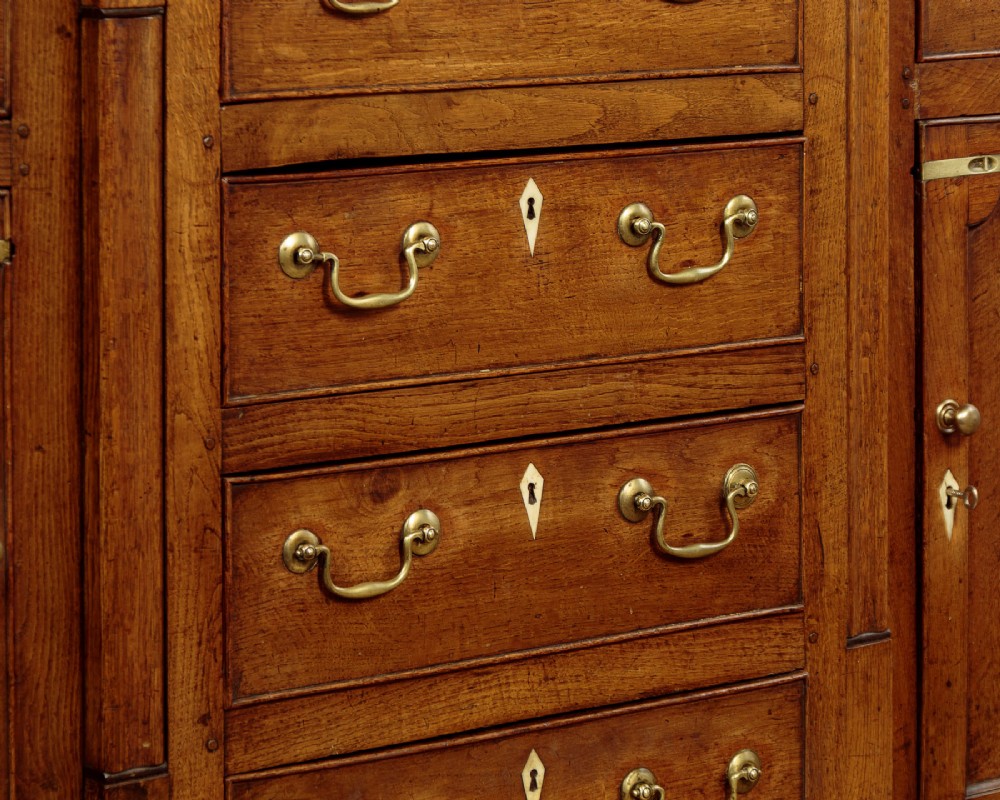 A late 18th-century warm brown oak Welsh dresser base featuring elevated top spice drawers, brass swan-neck handles, central drawers, and side cupboard doors. View X