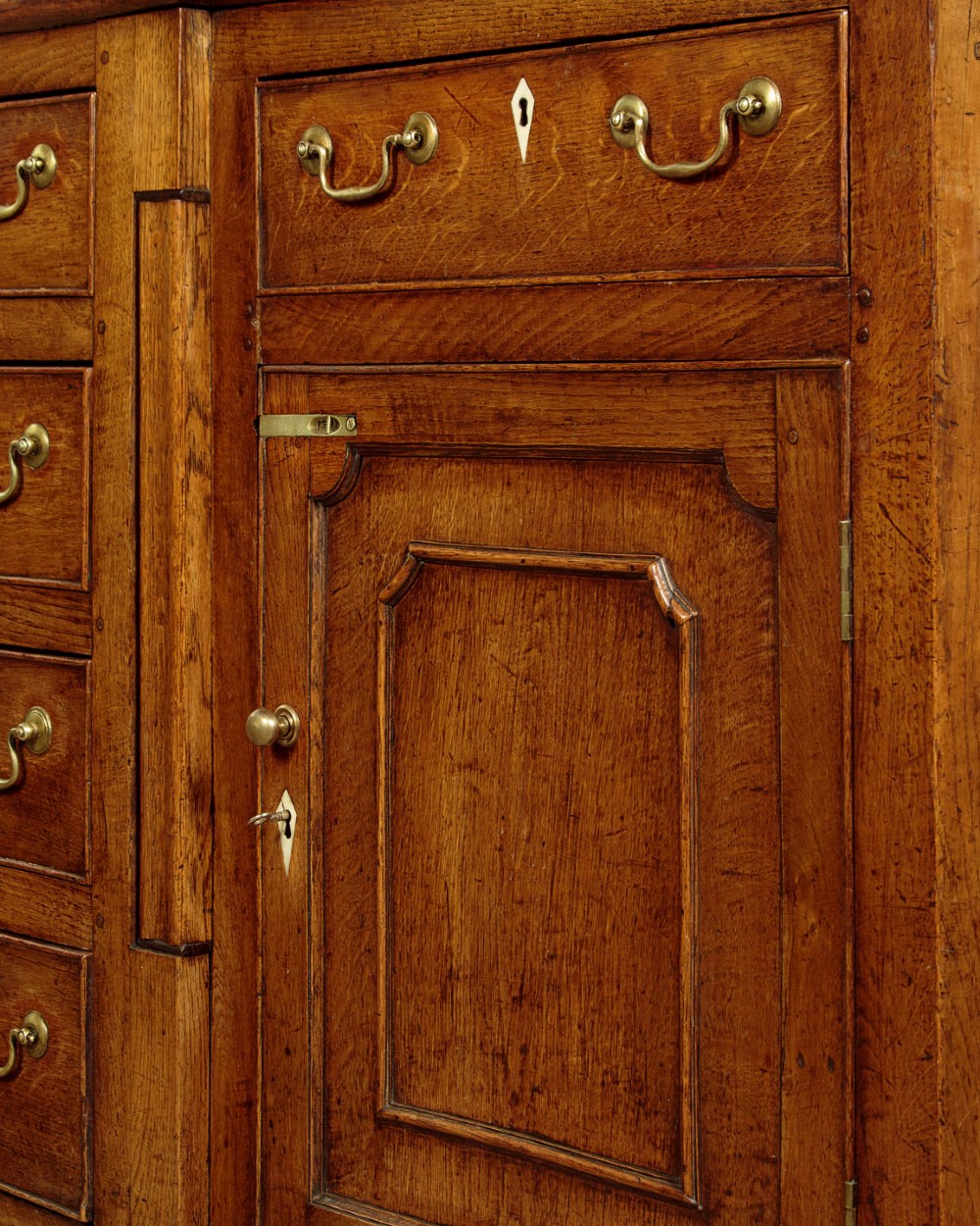 A late 18th-century warm brown oak Welsh dresser base featuring elevated top spice drawers, brass swan-neck handles, central drawers, and side cupboard doors. View XI