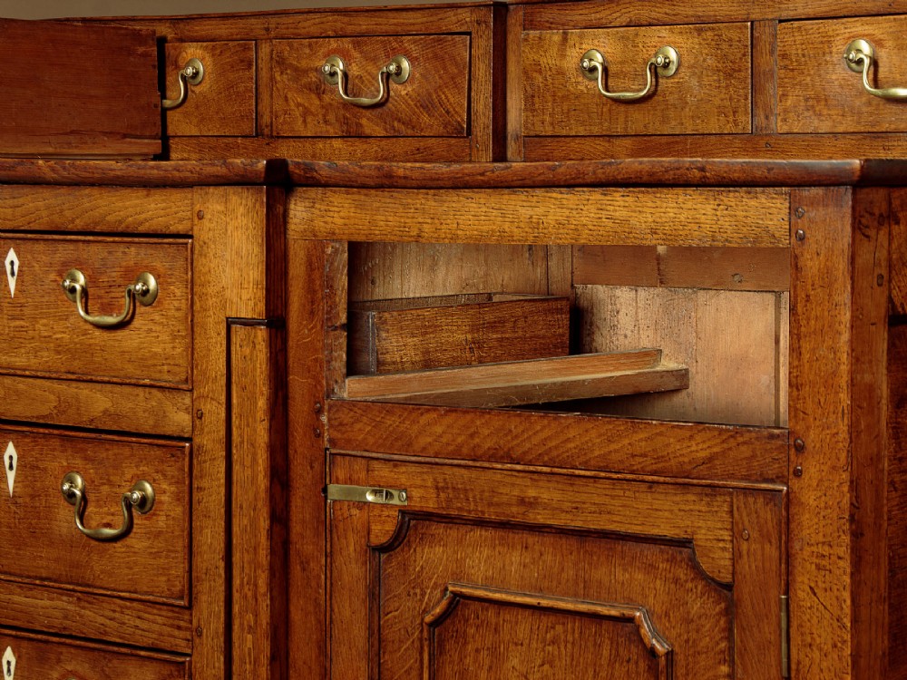 A late 18th-century warm brown oak Welsh dresser base featuring elevated top spice drawers, brass swan-neck handles, central drawers, and side cupboard doors. View XII