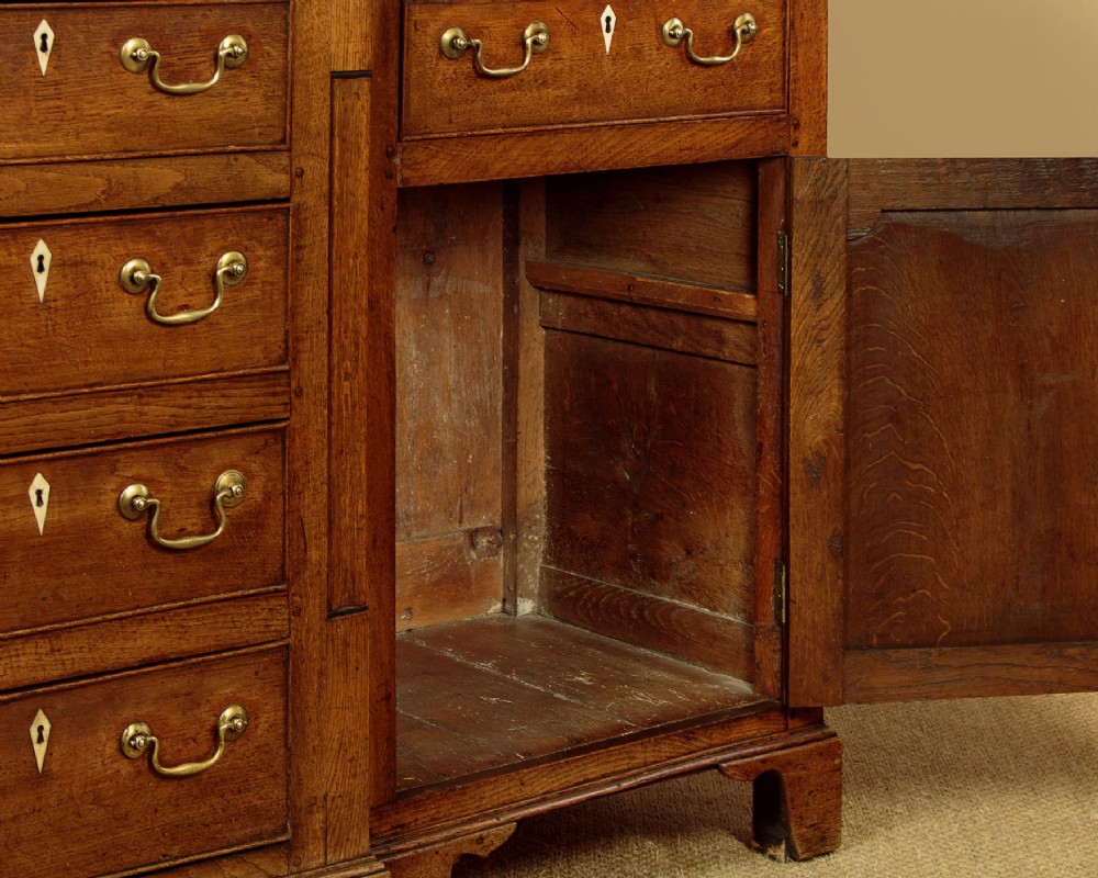 A late 18th-century warm brown oak Welsh dresser base featuring elevated top spice drawers, brass swan-neck handles, central drawers, and side cupboard doors. View XIV