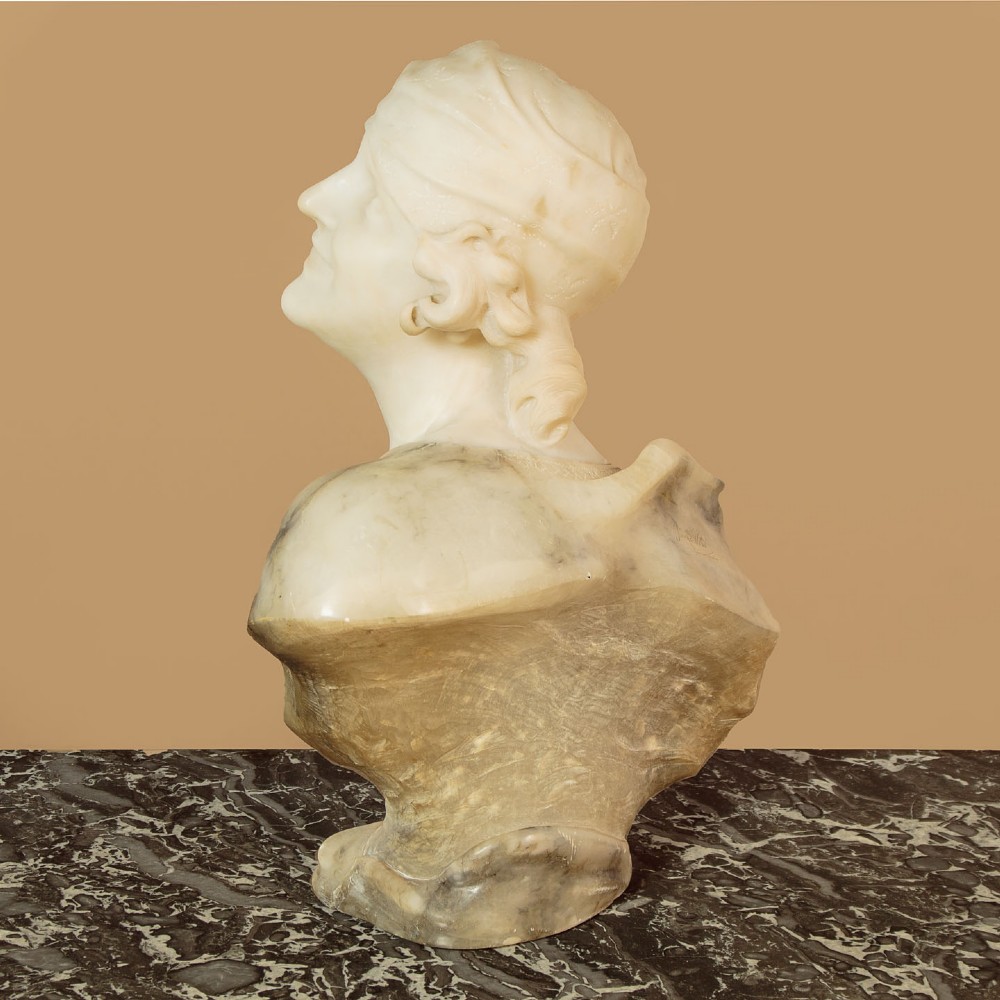 A carved antique alabaster bust of a woman wearing a headscarf tied with a bow, crafted from cream and veined stone. View X