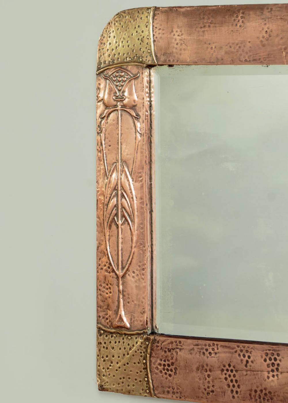 A rectangular Arts and Crafts wall mirror featuring a hand-hammered copper frame accented with repousse floral motifs and brass corner spandrels. View V