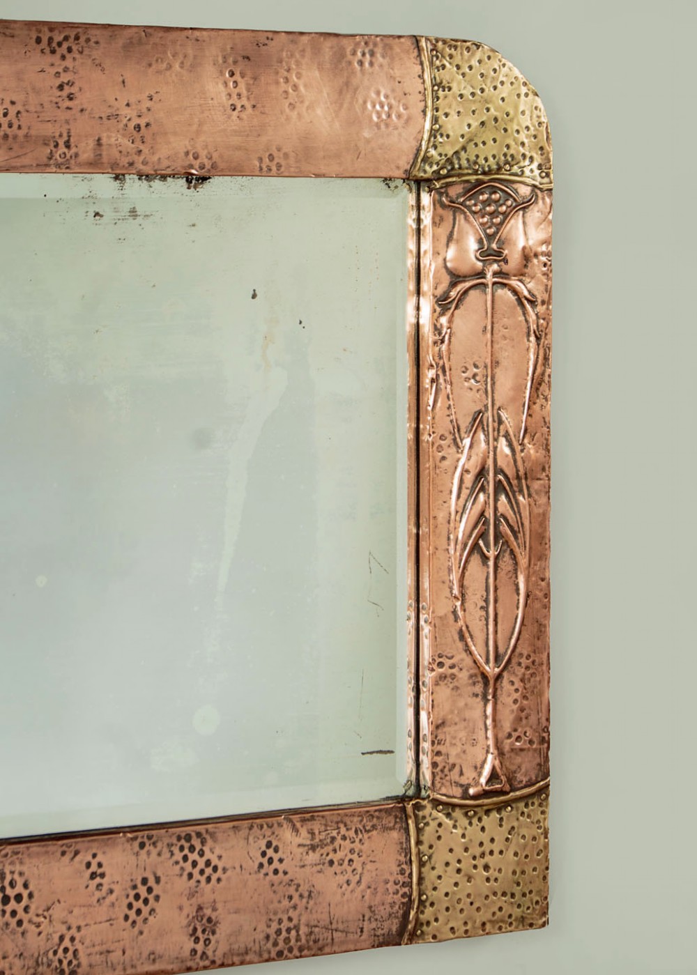 A rectangular Arts and Crafts wall mirror featuring a hand-hammered copper frame accented with repousse floral motifs and brass corner spandrels. View VI