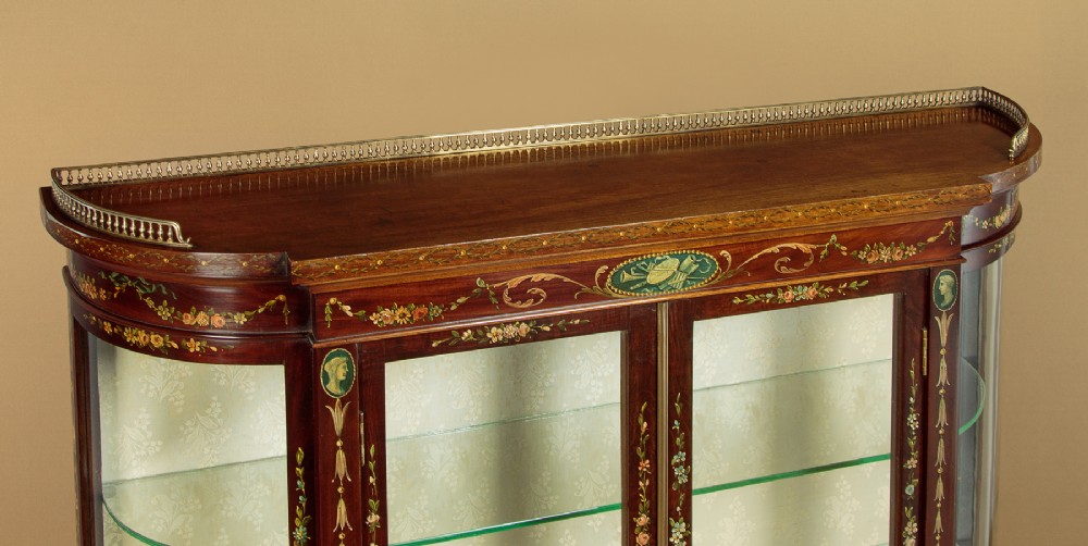 A dark mahogany display cabinet featuring hand-painted floral swags, classical urns, glass side panels, twin glazed doors, and tapered legs with an undertier. View IV