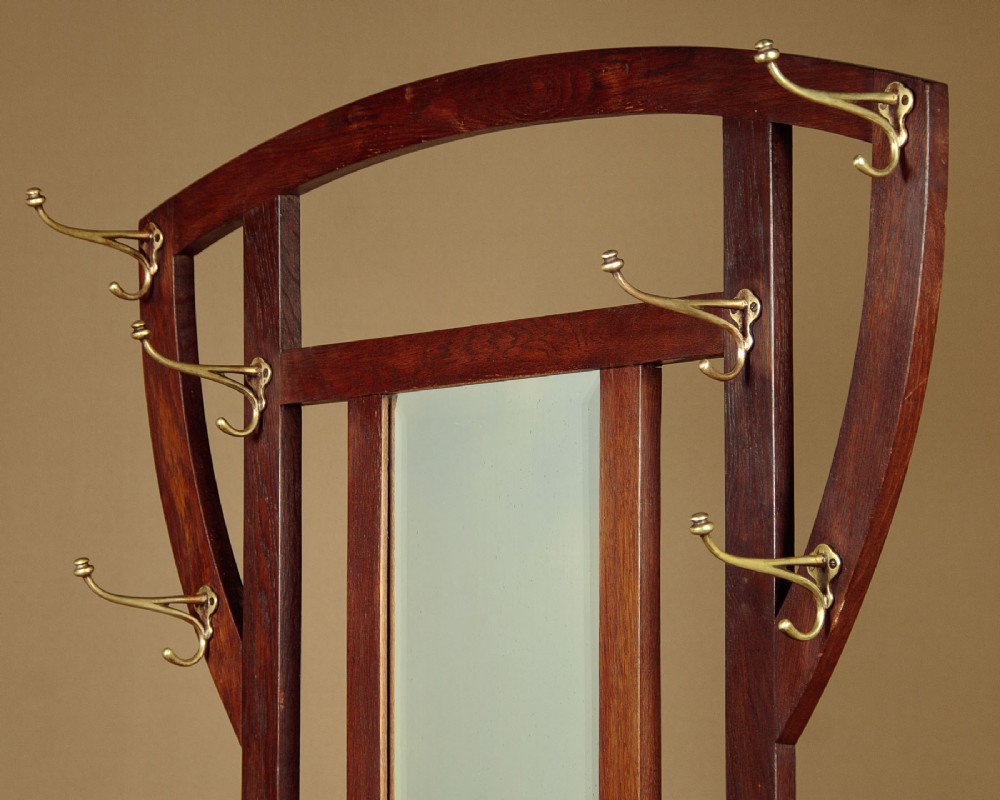 An Art Nouveau oak hallstand featuring an arched upper frame, brass coat hooks, central mirror, glove box, and bottom umbrella stand with two brass drip trays. View III