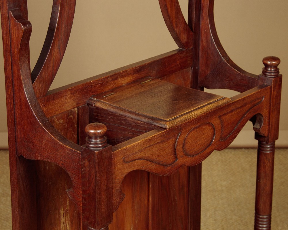 An Art Nouveau oak hallstand featuring an arched upper frame, brass coat hooks, central mirror, glove box, and bottom umbrella stand with two brass drip trays. View VII