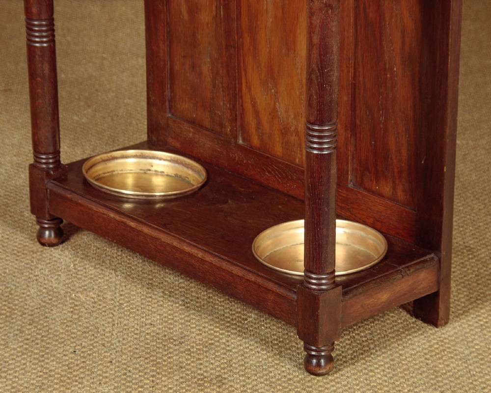 An Art Nouveau oak hallstand featuring an arched upper frame, brass coat hooks, central mirror, glove box, and bottom umbrella stand with two brass drip trays. View X