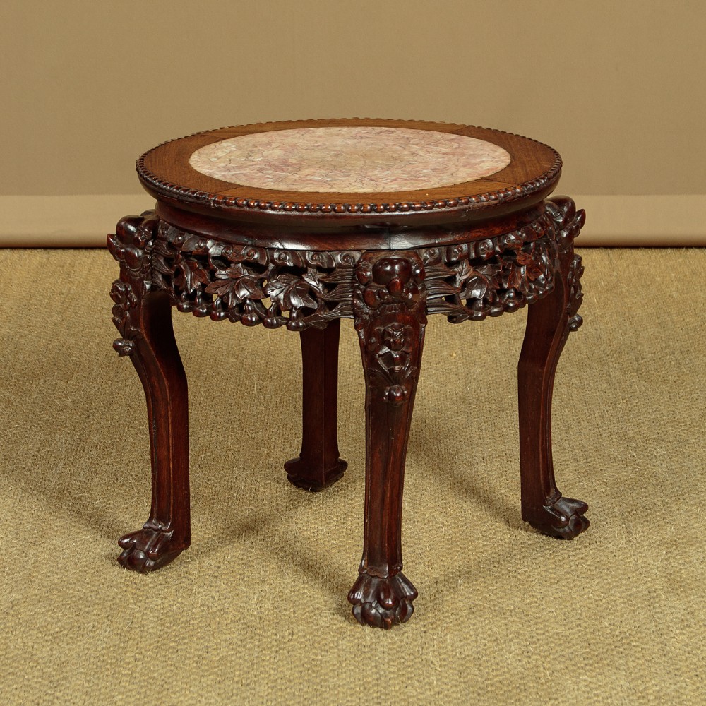 A low circular dark hardwood side table featuring an inset circular pink marble top, deeply carved floral aprons, and cabriole legs terminating in claw feet.