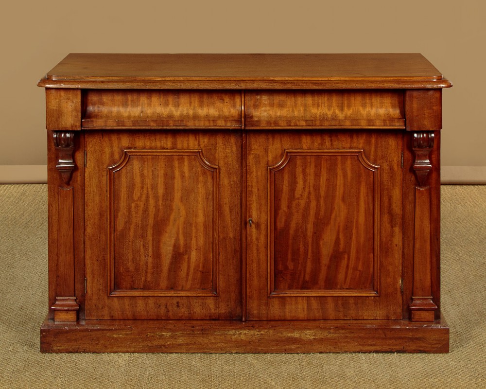 A mid-19th-century Victorian mahogany sideboard featuring a smooth flat top, two frieze drawers, and a pair of panelled cupboard doors flanked by carved corbels.
