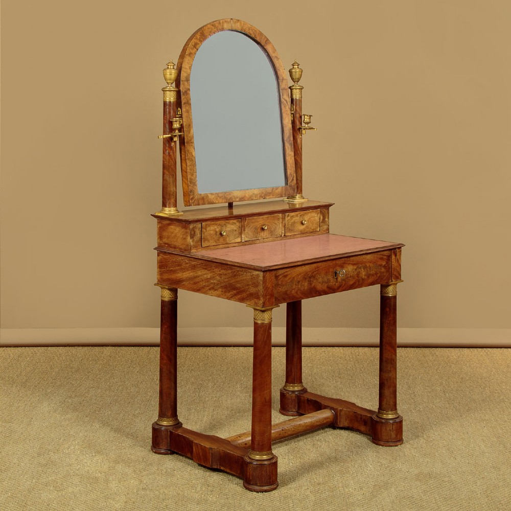 A French Empire flame mahogany dressing table featuring an arched swing mirror flanked by column supports with brass mounts, small trinket drawers, and a pink leather writing surface.