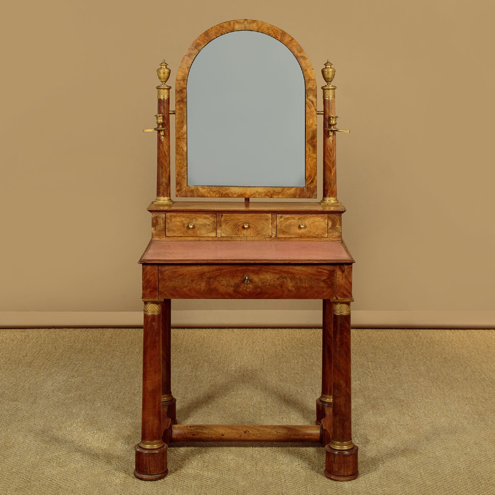 A French Empire flame mahogany dressing table featuring an arched swing mirror flanked by column supports with brass mounts, small trinket drawers, and a pink leather writing surface. View II