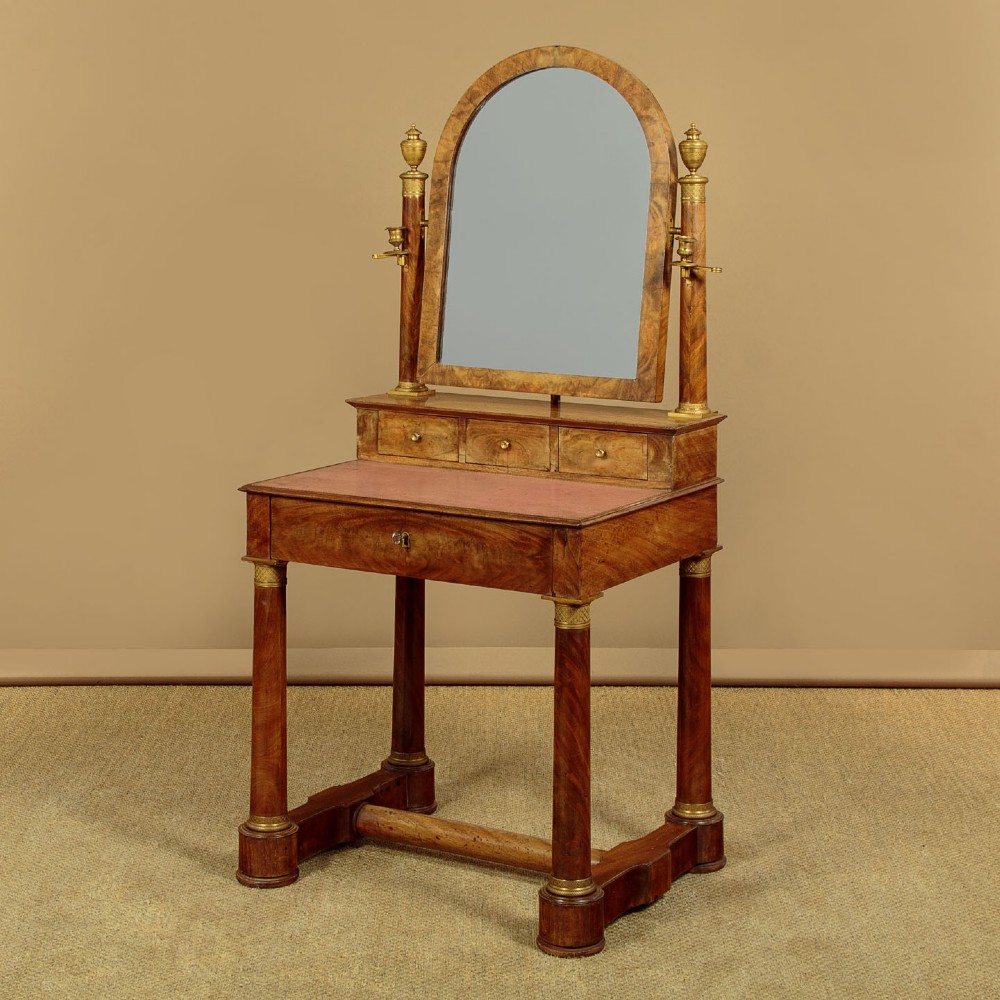 A French Empire flame mahogany dressing table featuring an arched swing mirror flanked by column supports with brass mounts, small trinket drawers, and a pink leather writing surface. View III