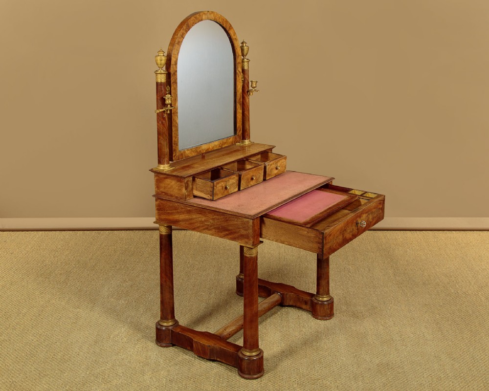 A French Empire flame mahogany dressing table featuring an arched swing mirror flanked by column supports with brass mounts, small trinket drawers, and a pink leather writing surface. View IV