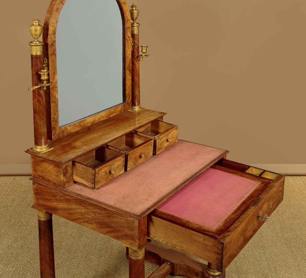 A French Empire flame mahogany dressing table featuring an arched swing mirror flanked by column supports with brass mounts, small trinket drawers, and a pink leather writing surface. View VI