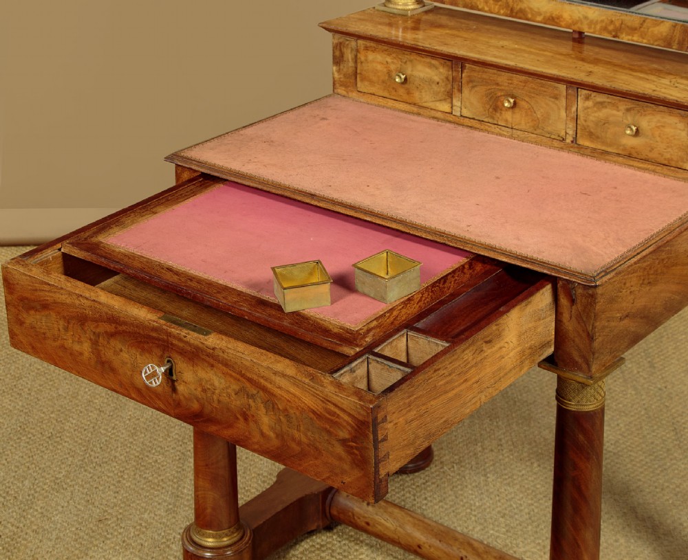 A French Empire flame mahogany dressing table featuring an arched swing mirror flanked by column supports with brass mounts, small trinket drawers, and a pink leather writing surface. View VIII