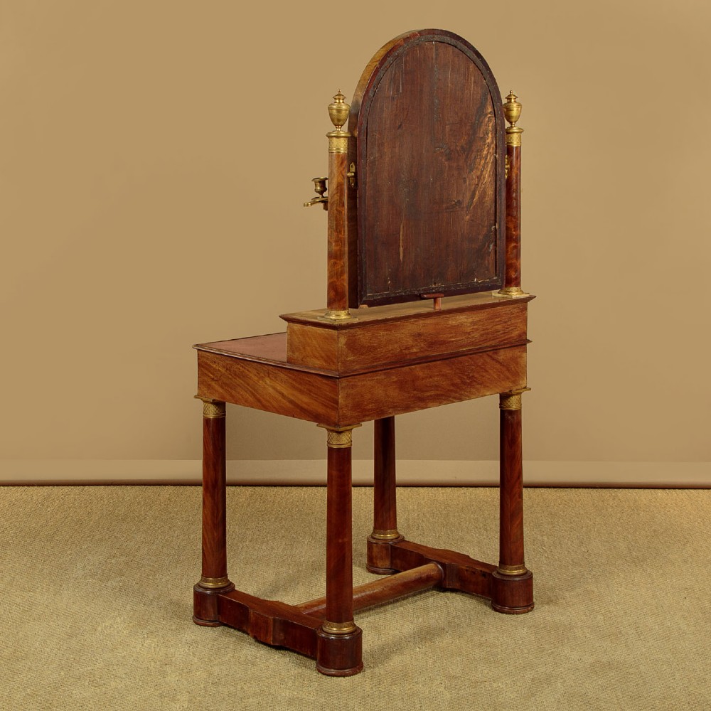 A French Empire flame mahogany dressing table featuring an arched swing mirror flanked by column supports with brass mounts, small trinket drawers, and a pink leather writing surface. View XI