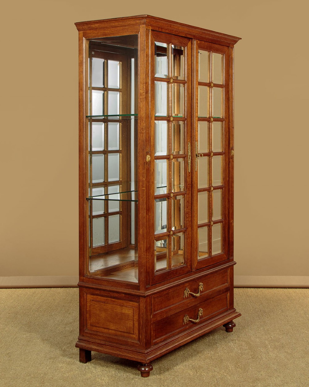 An Edwardian oak display cabinet featuring two glazed doors with gilt rosette mounts, a mirrored interior back, and two drawers at the base with swag handles. View II