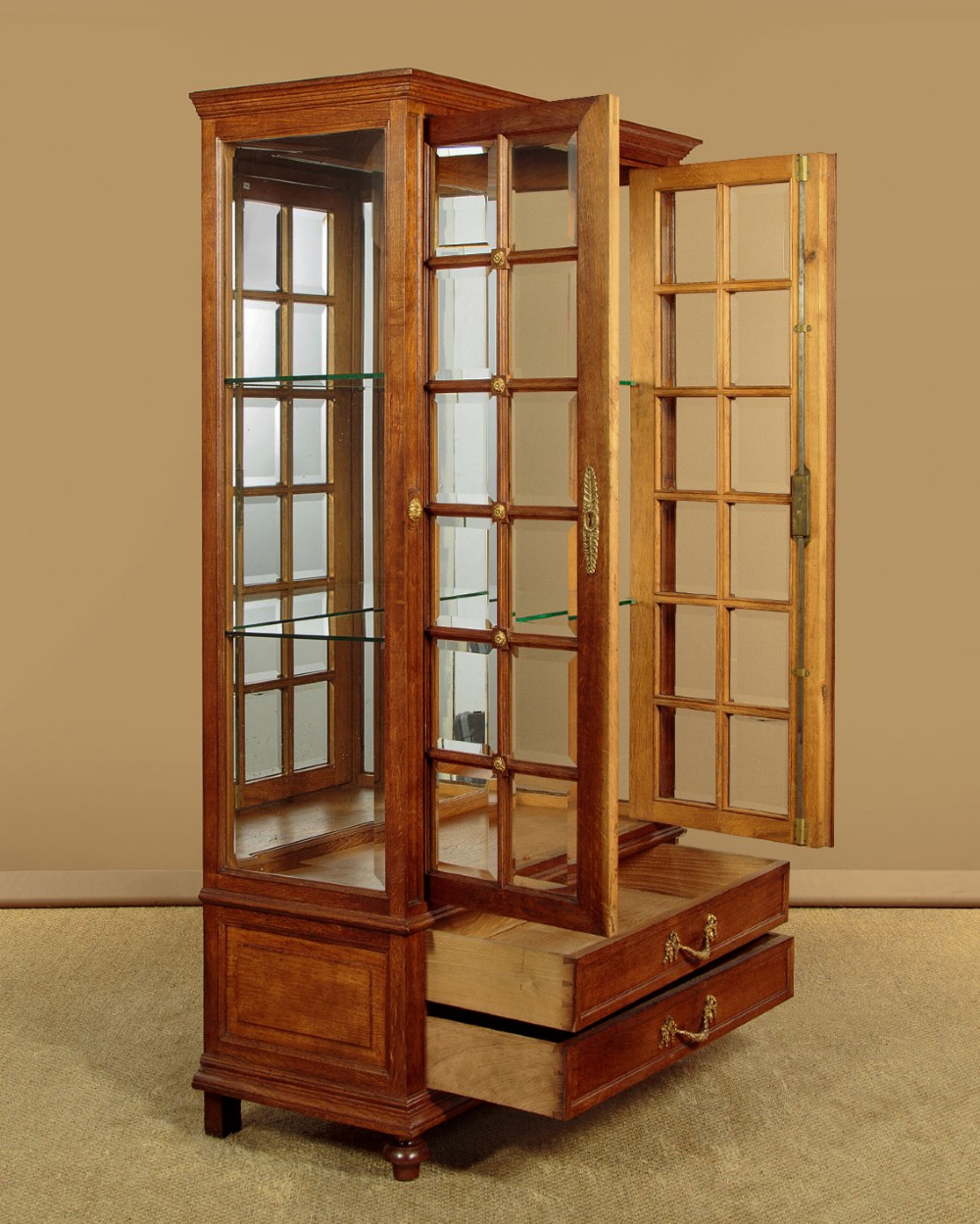 An Edwardian oak display cabinet featuring two glazed doors with gilt rosette mounts, a mirrored interior back, and two drawers at the base with swag handles. View IV
