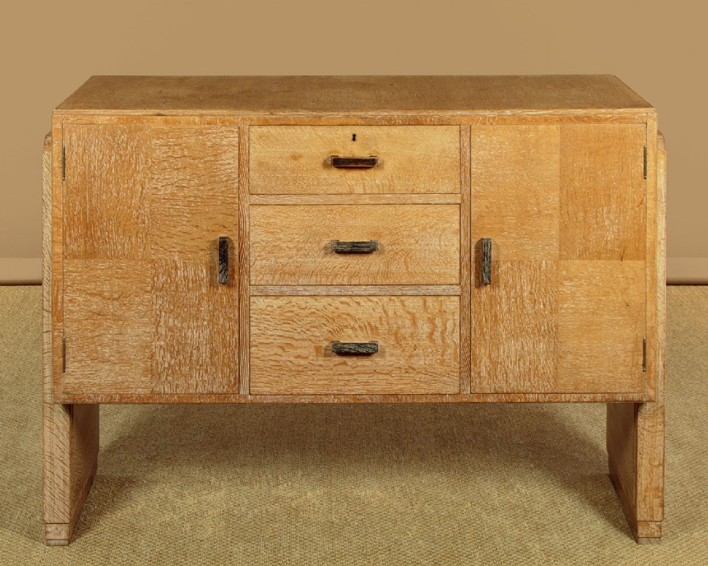 A 1920s limed oak sideboard featuring a central bank of three drawers flanked by two panelled cupboard doors on slab feet.