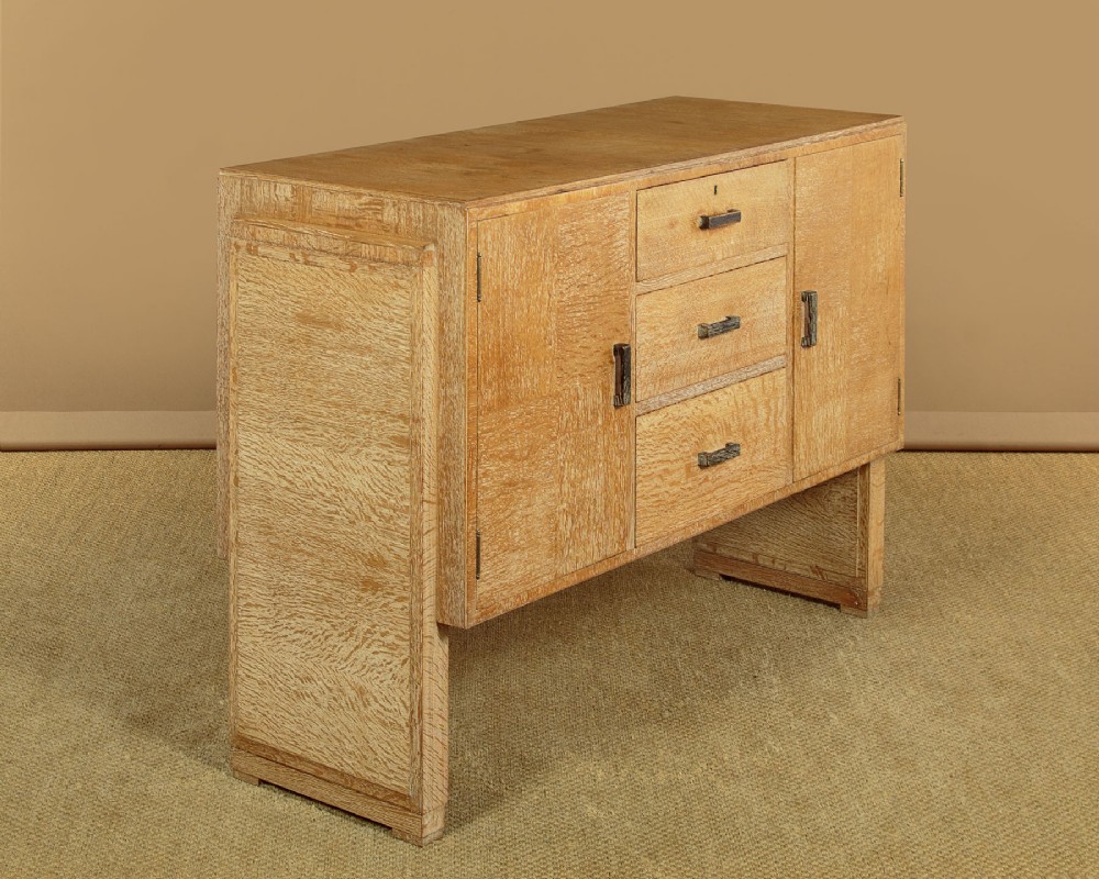 A 1920s limed oak sideboard featuring a central bank of three drawers flanked by two panelled cupboard doors on slab feet. View II