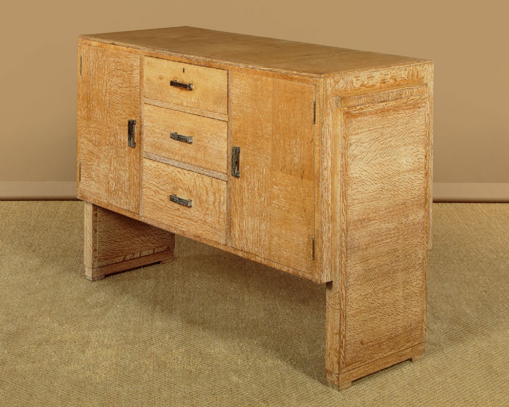 A 1920s limed oak sideboard featuring a central bank of three drawers flanked by two panelled cupboard doors on slab feet. View III