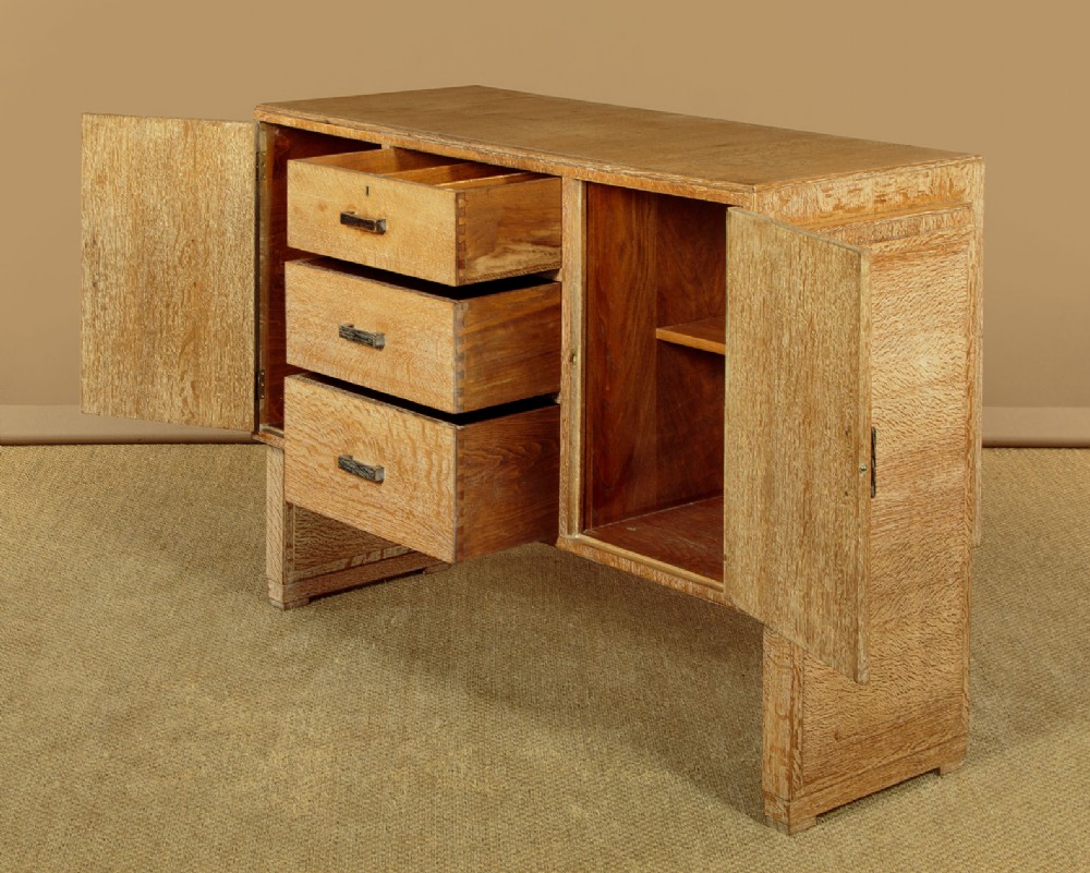 A 1920s limed oak sideboard featuring a central bank of three drawers flanked by two panelled cupboard doors on slab feet. View V