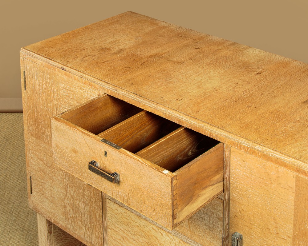 A 1920s limed oak sideboard featuring a central bank of three drawers flanked by two panelled cupboard doors on slab feet. View VII