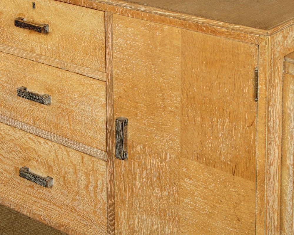 A 1920s limed oak sideboard featuring a central bank of three drawers flanked by two panelled cupboard doors on slab feet. View XI