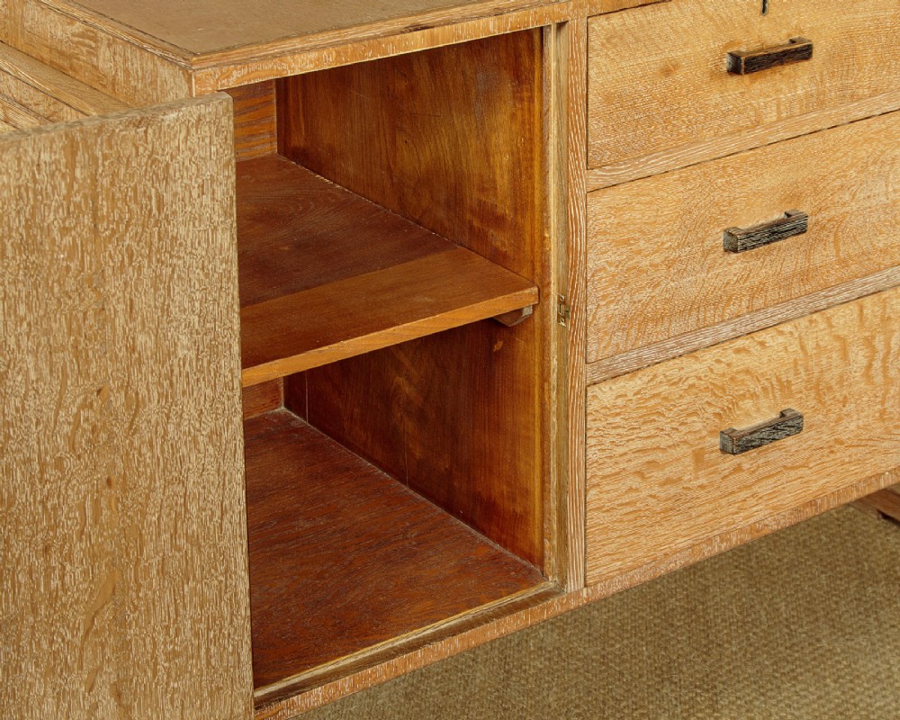 A 1920s limed oak sideboard featuring a central bank of three drawers flanked by two panelled cupboard doors on slab feet. View XII