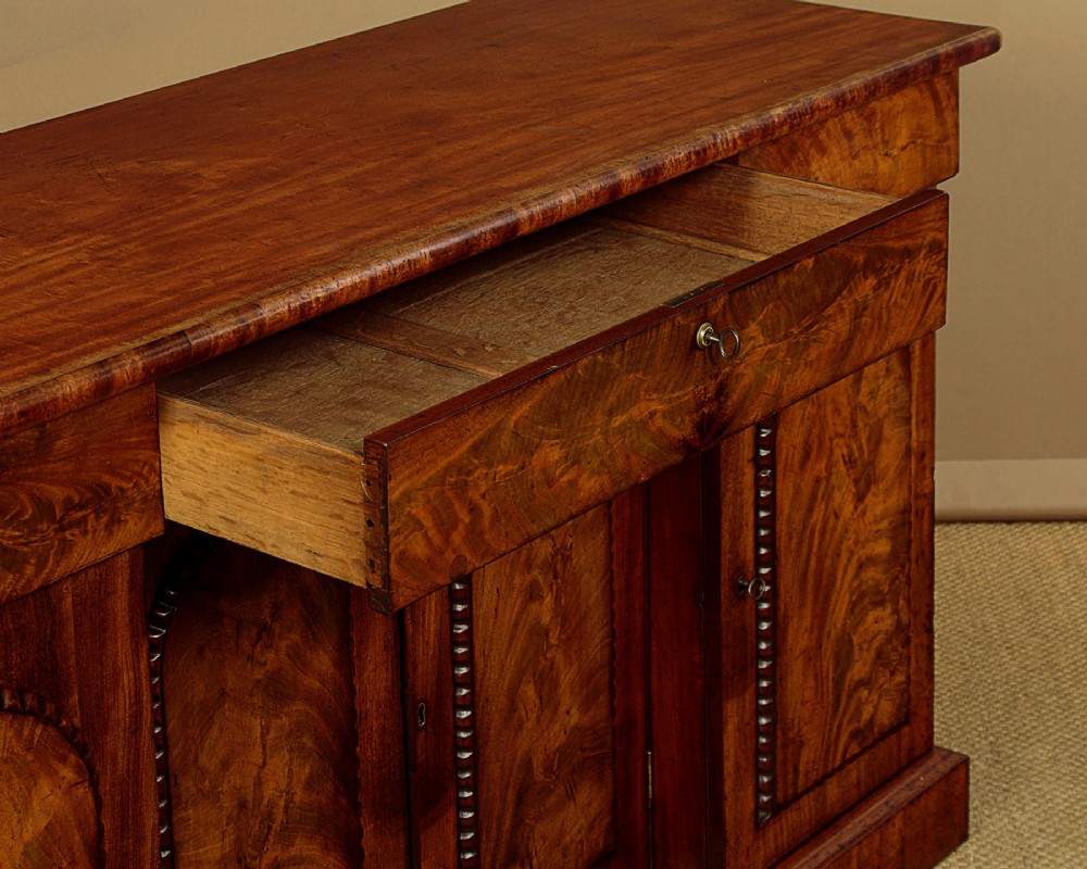 A William IV mahogany four-door breakfront sideboard featuring flame mahogany drawer fronts and arched cupboard doors set on a solid plinth base. View VI