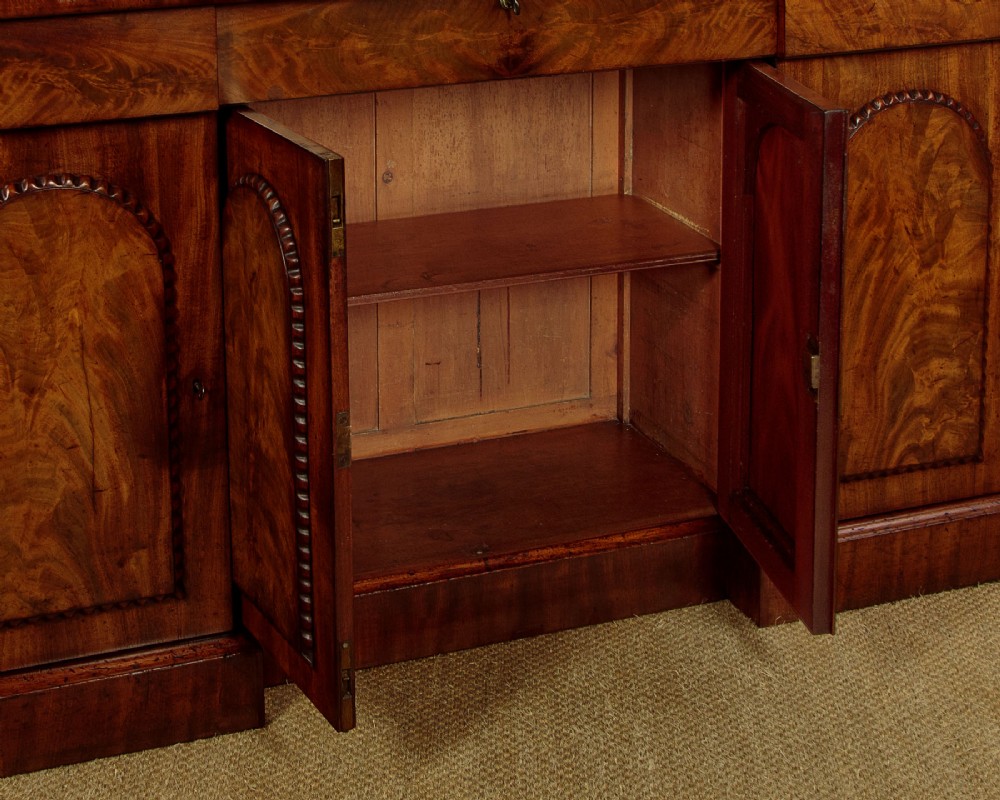 A William IV mahogany four-door breakfront sideboard featuring flame mahogany drawer fronts and arched cupboard doors set on a solid plinth base. View X