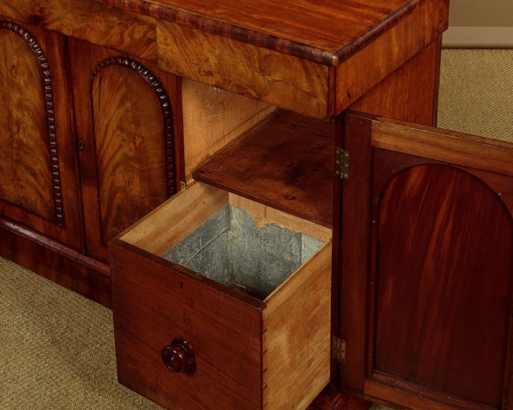 A William IV mahogany four-door breakfront sideboard featuring flame mahogany drawer fronts and arched cupboard doors set on a solid plinth base. View XI
