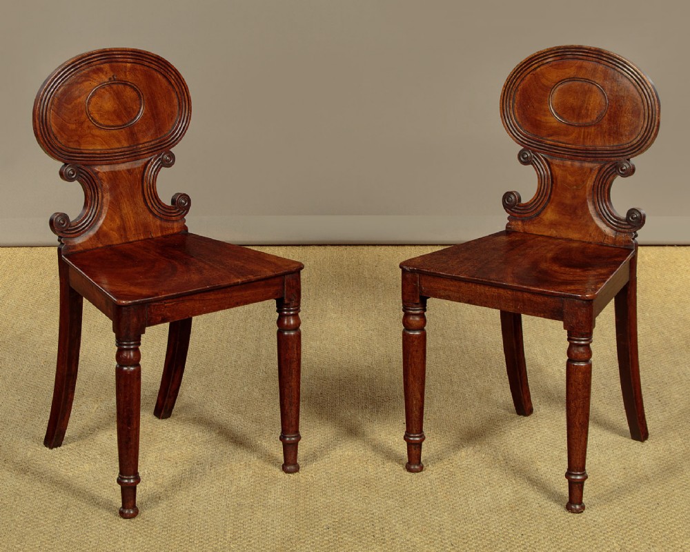 A pair of early nineteenth-century Regency mahogany hall chairs with carved oval backrests, scroll waist accents, solid wooden seats, and turned front legs.