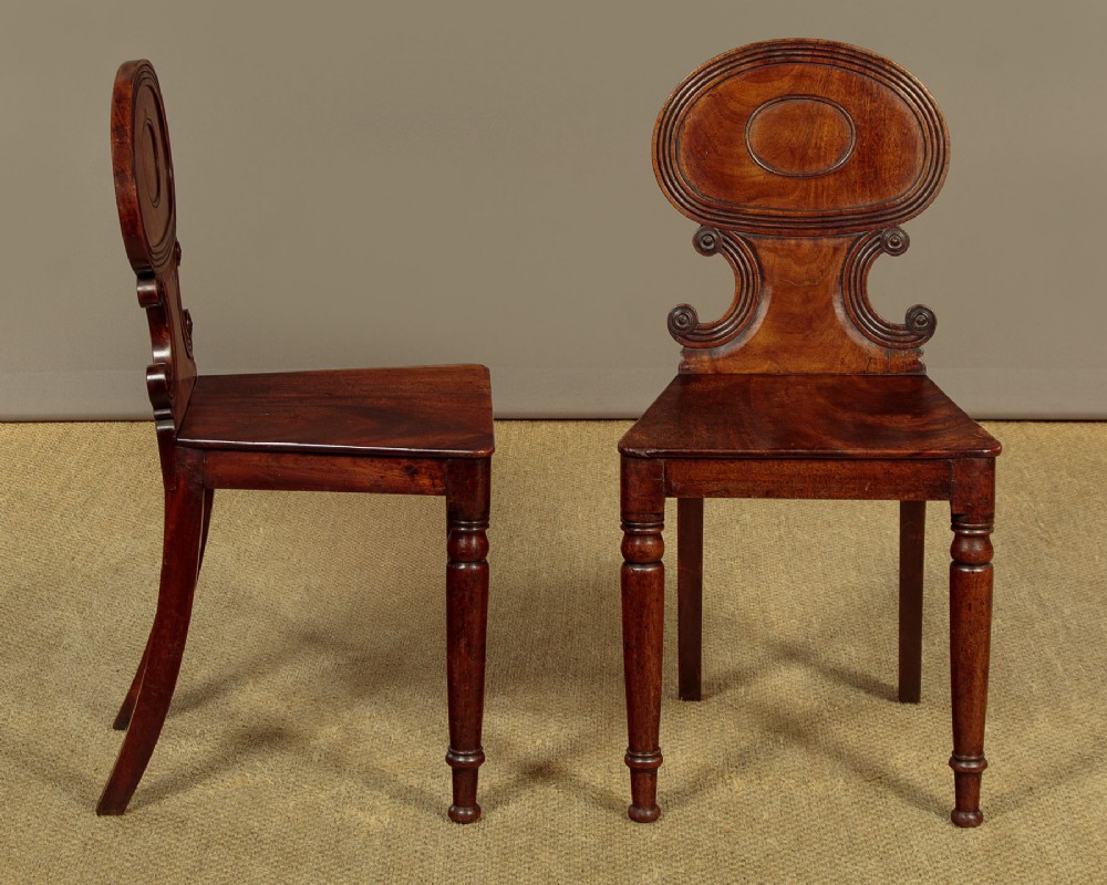 A pair of early nineteenth-century Regency mahogany hall chairs with carved oval backrests, scroll waist accents, solid wooden seats, and turned front legs. View II