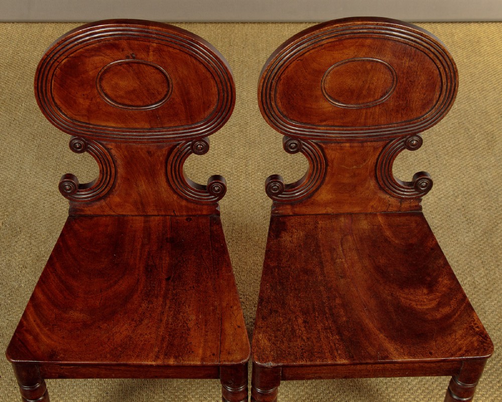 A pair of early nineteenth-century Regency mahogany hall chairs with carved oval backrests, scroll waist accents, solid wooden seats, and turned front legs. View III