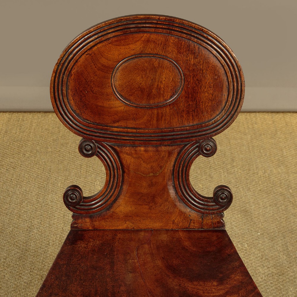 A pair of early nineteenth-century Regency mahogany hall chairs with carved oval backrests, scroll waist accents, solid wooden seats, and turned front legs. View IV