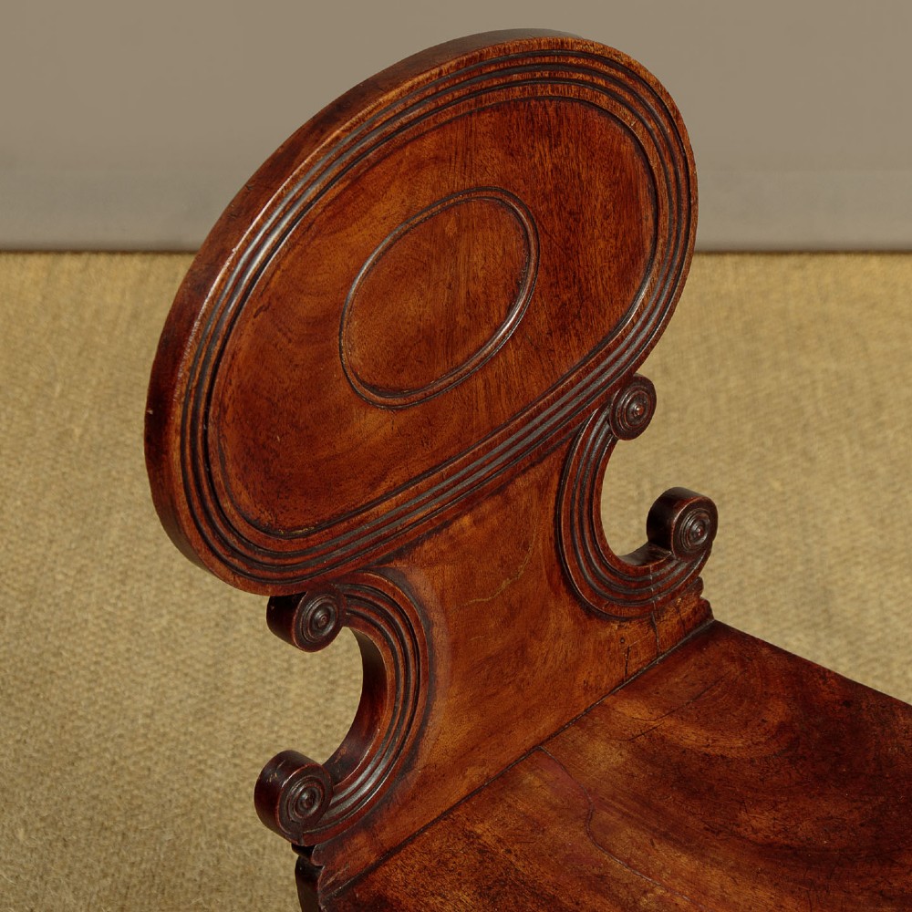 A pair of early nineteenth-century Regency mahogany hall chairs with carved oval backrests, scroll waist accents, solid wooden seats, and turned front legs. View V