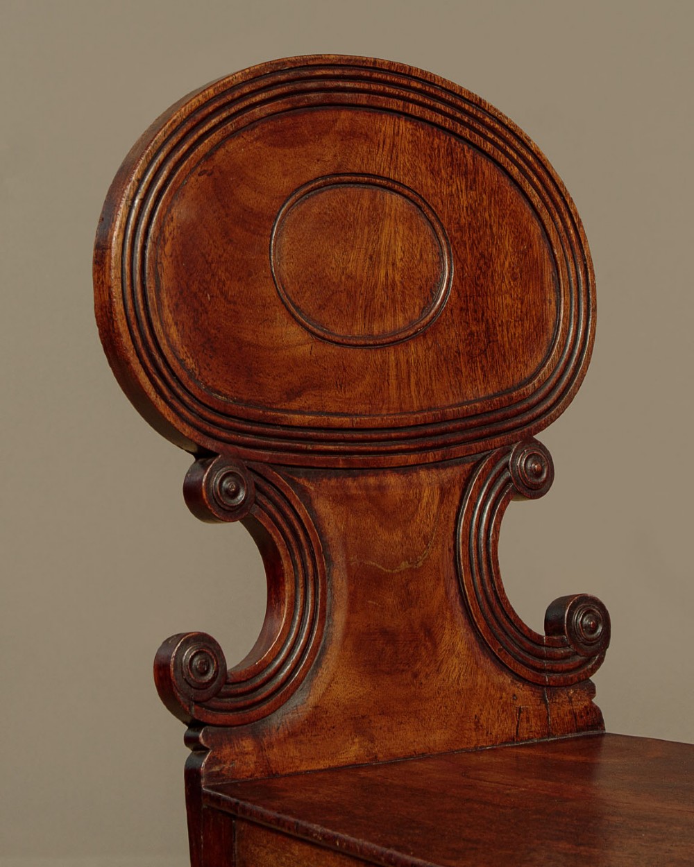 A pair of early nineteenth-century Regency mahogany hall chairs with carved oval backrests, scroll waist accents, solid wooden seats, and turned front legs. View VI
